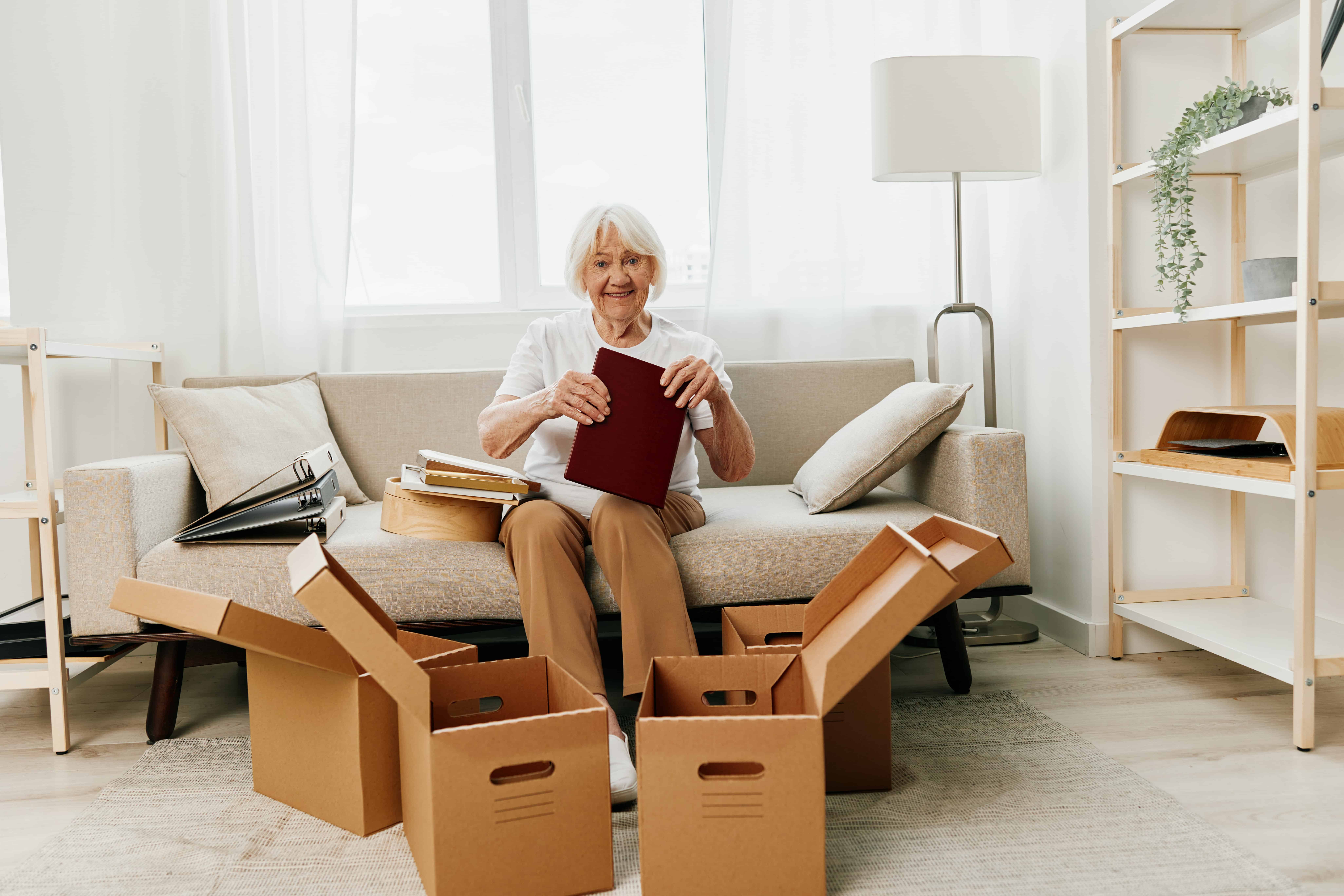 elderly woman sits on a sofa at home with boxes. collecting things with memories and moving and happiness smile. Woman packing